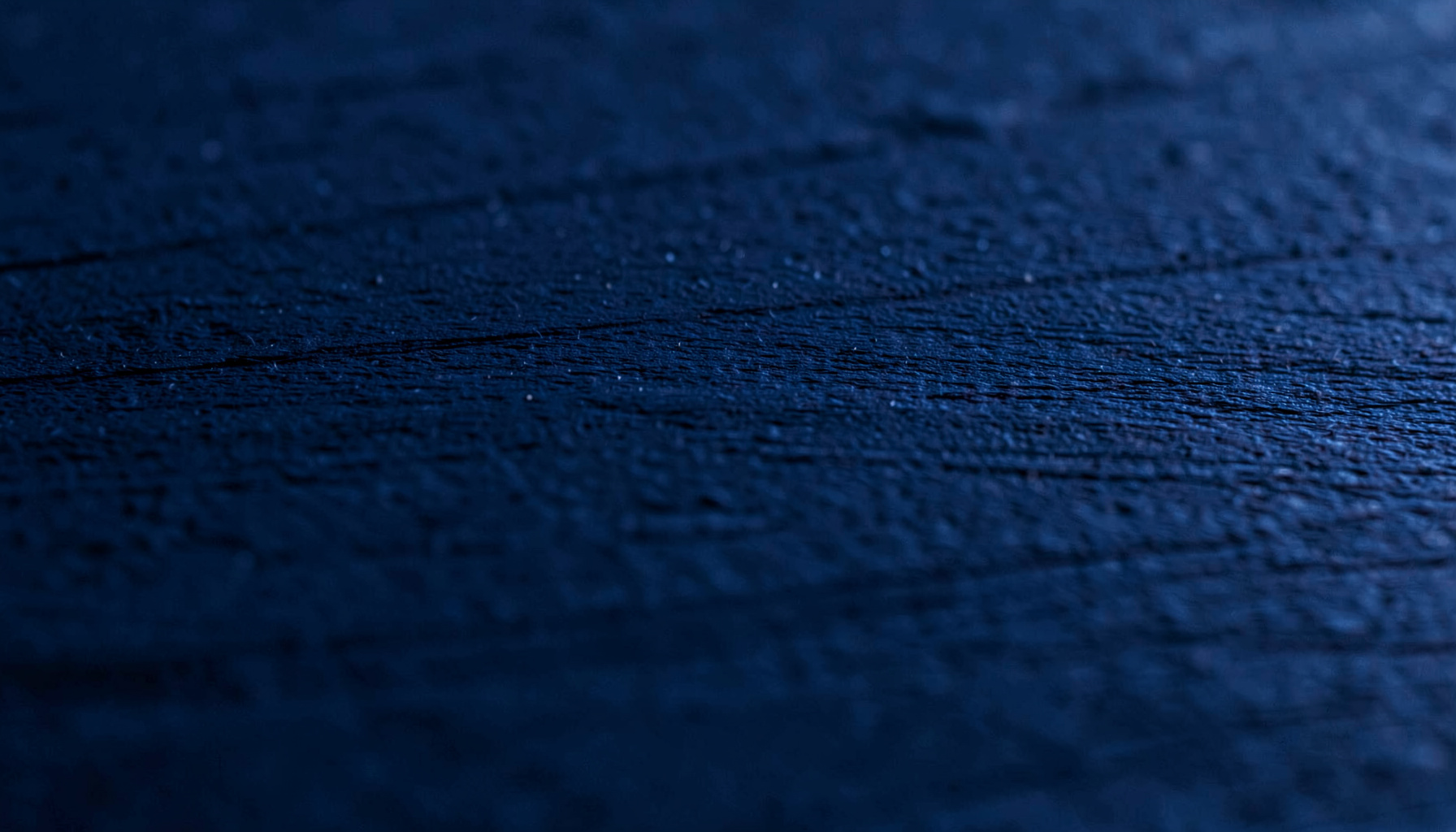 A close-up photograph of a dark navy-blue textured surface with subtle horizontal grain, moody and understated.